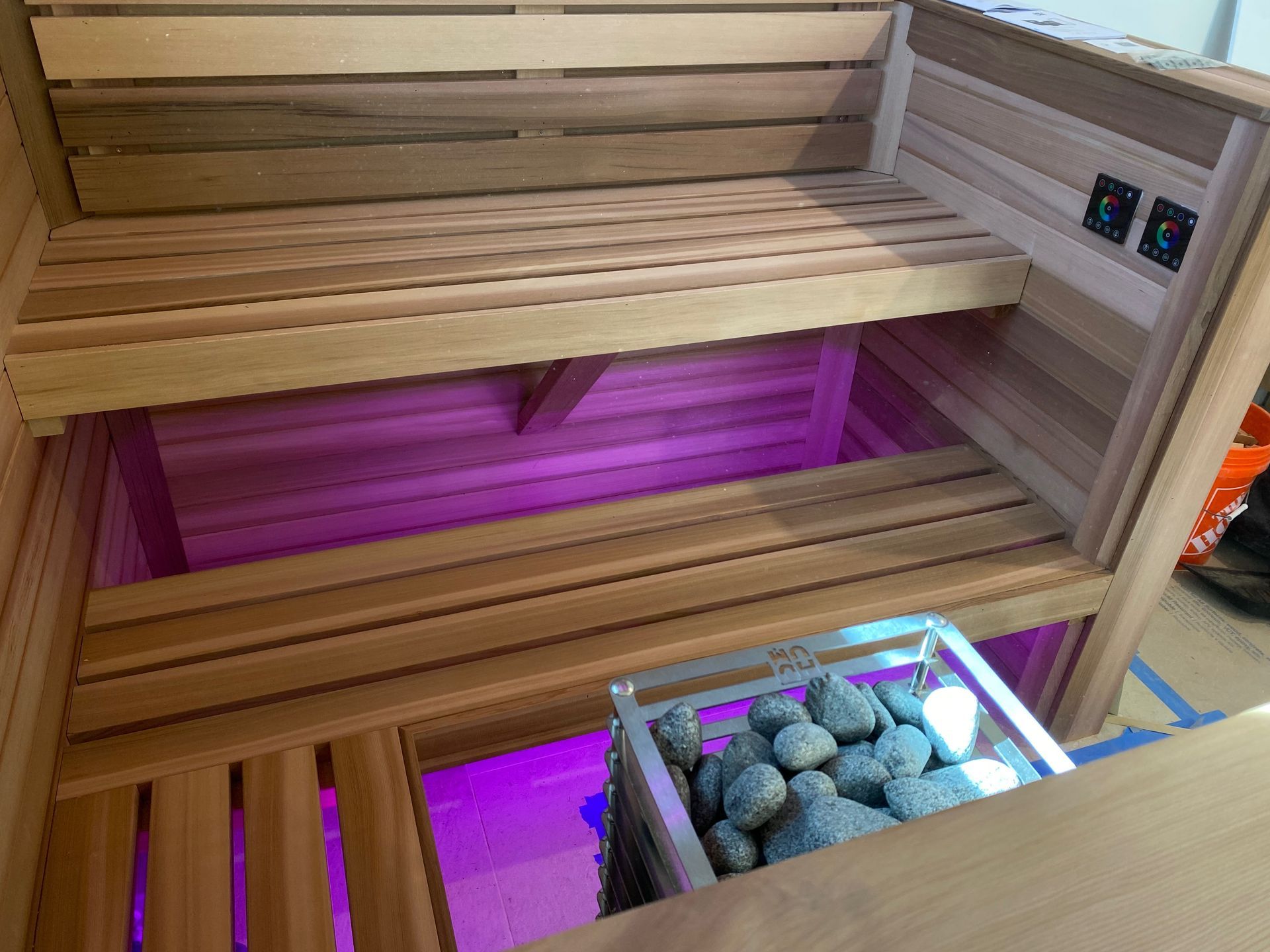 Wooden sauna interior with benches, purple lighting, and a rock-filled heater.