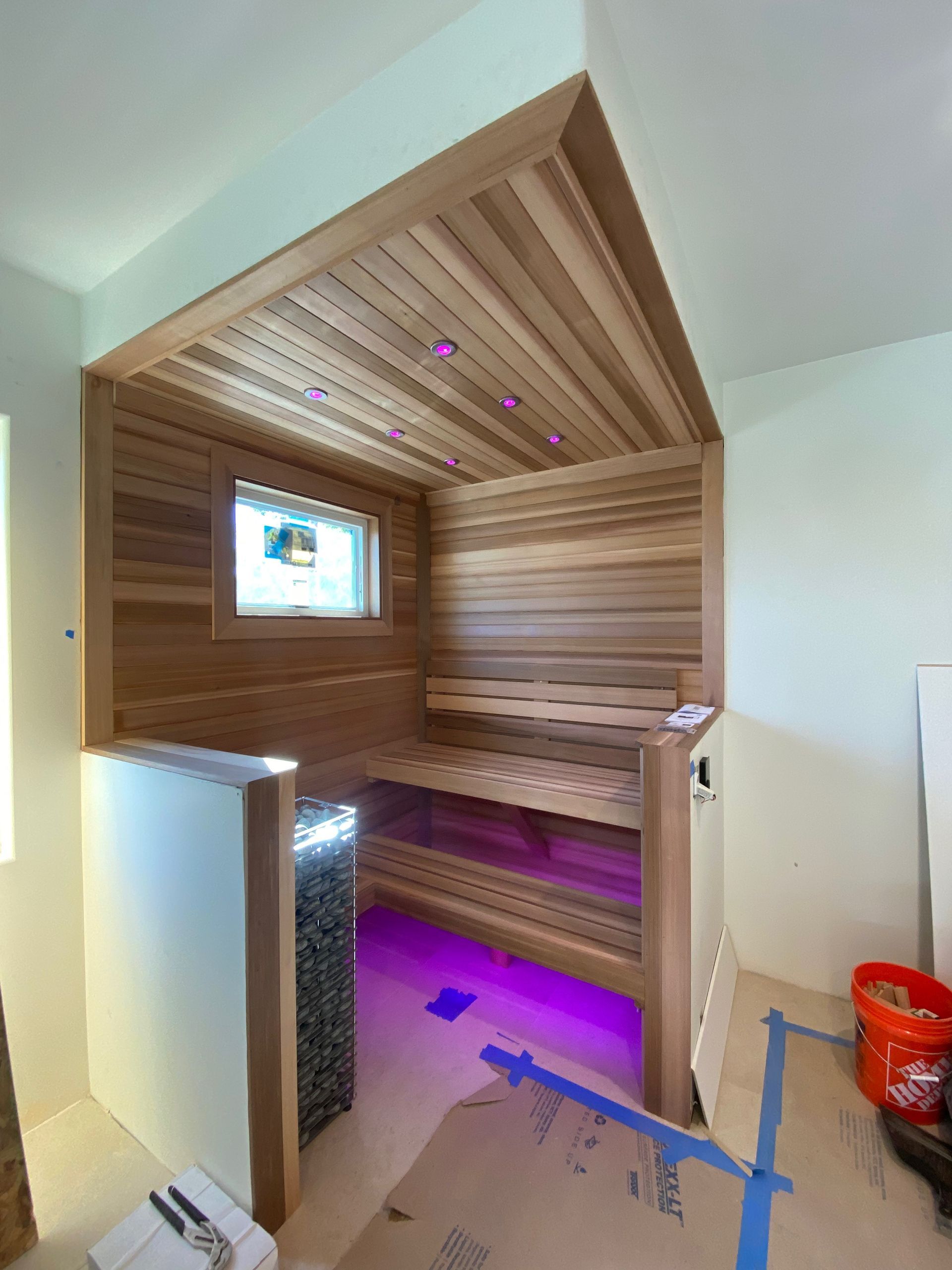 Wooden sauna with bench seating, ceiling lights, and small window. Purple under-bench lighting.