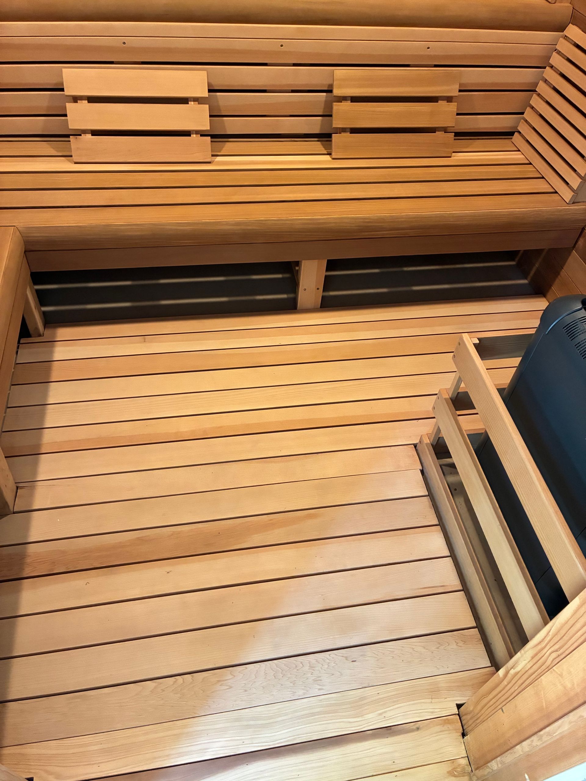 Interior of a wooden sauna with bench seating, slatted flooring, and a heater in the corner.