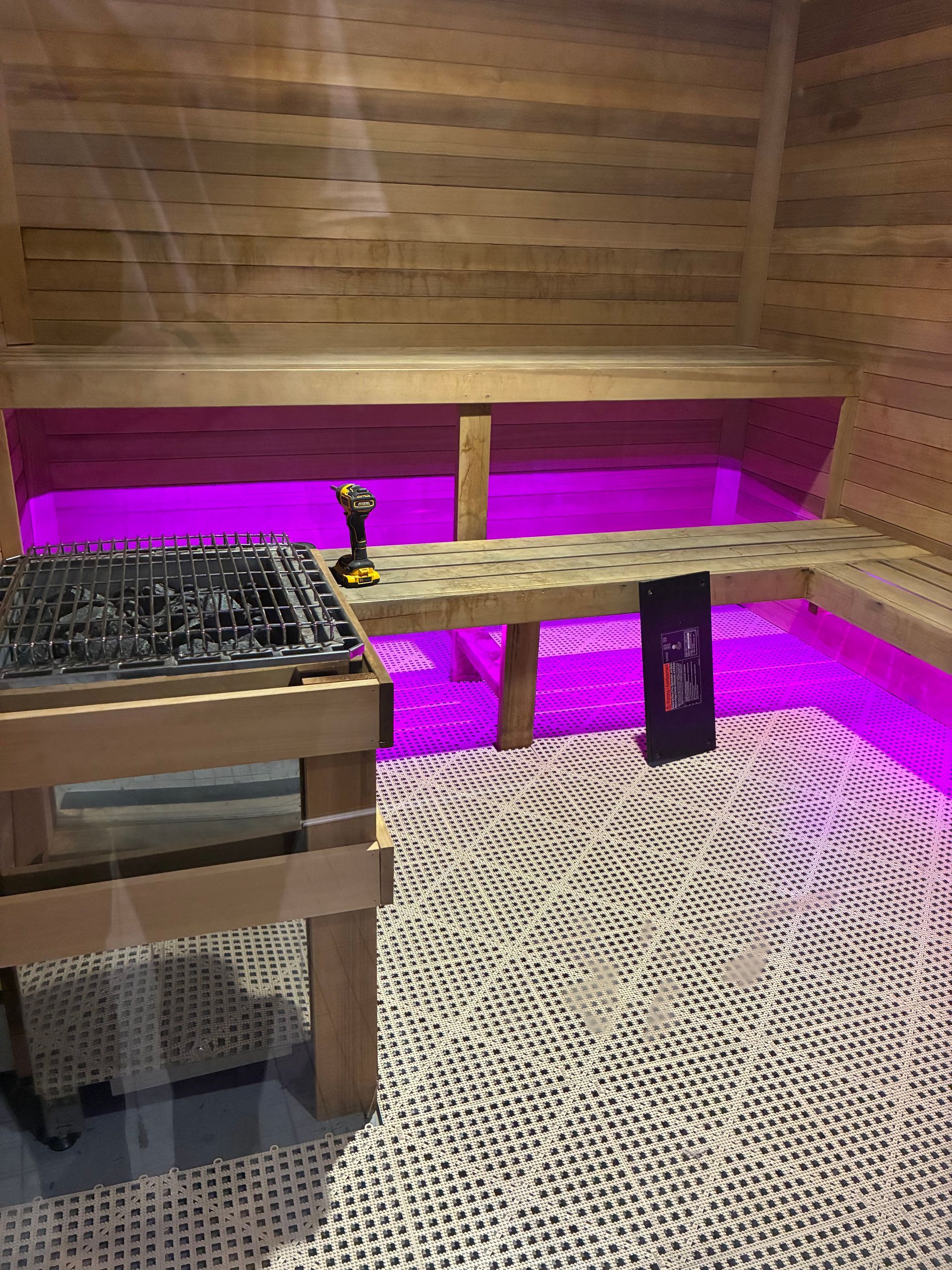 Interior of a sauna with wooden benches, a heater, and purple lighting. The floor has a white, textured mat.