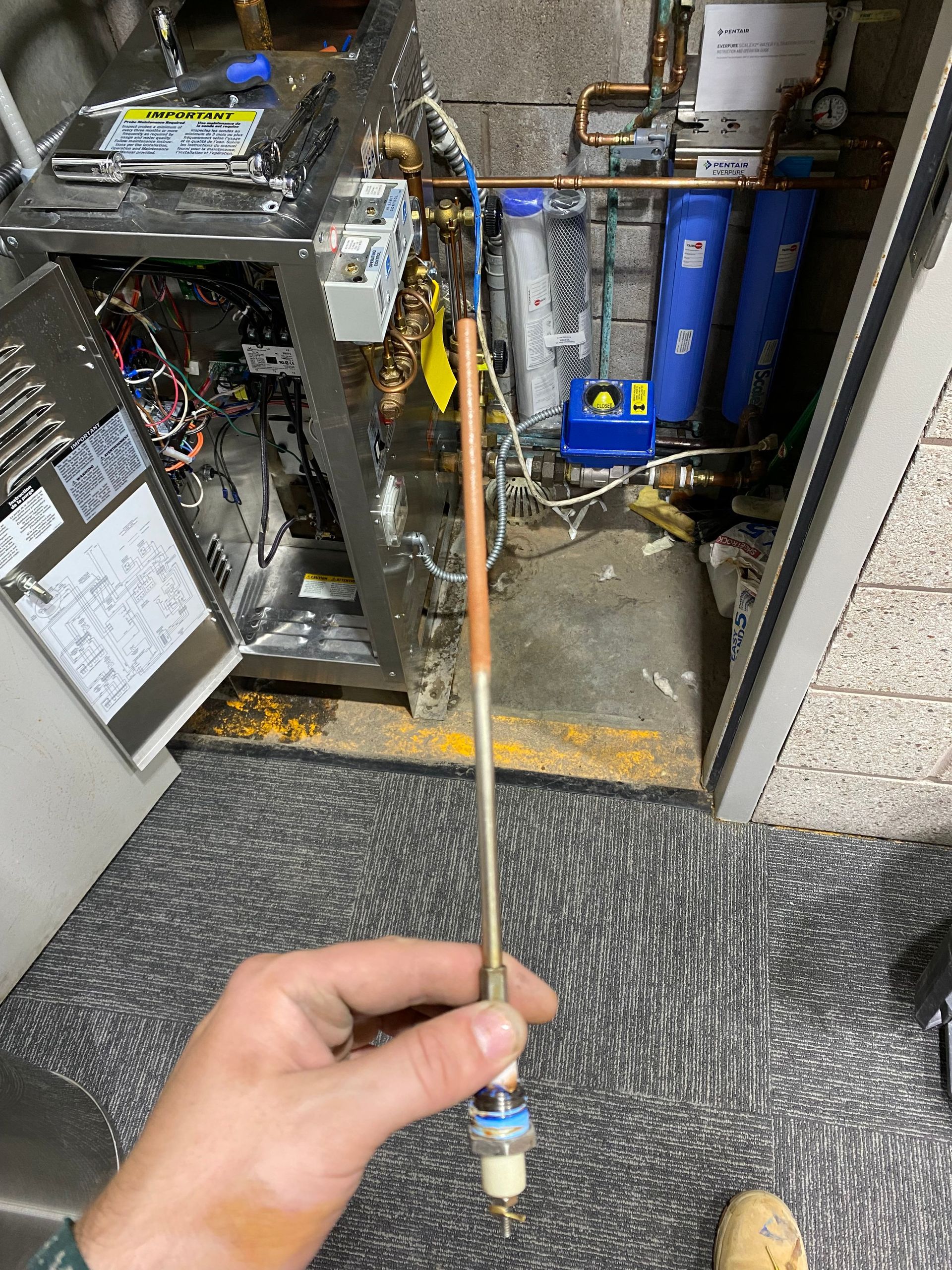 Hand holding a long, slender metal probe; heating system components in background.