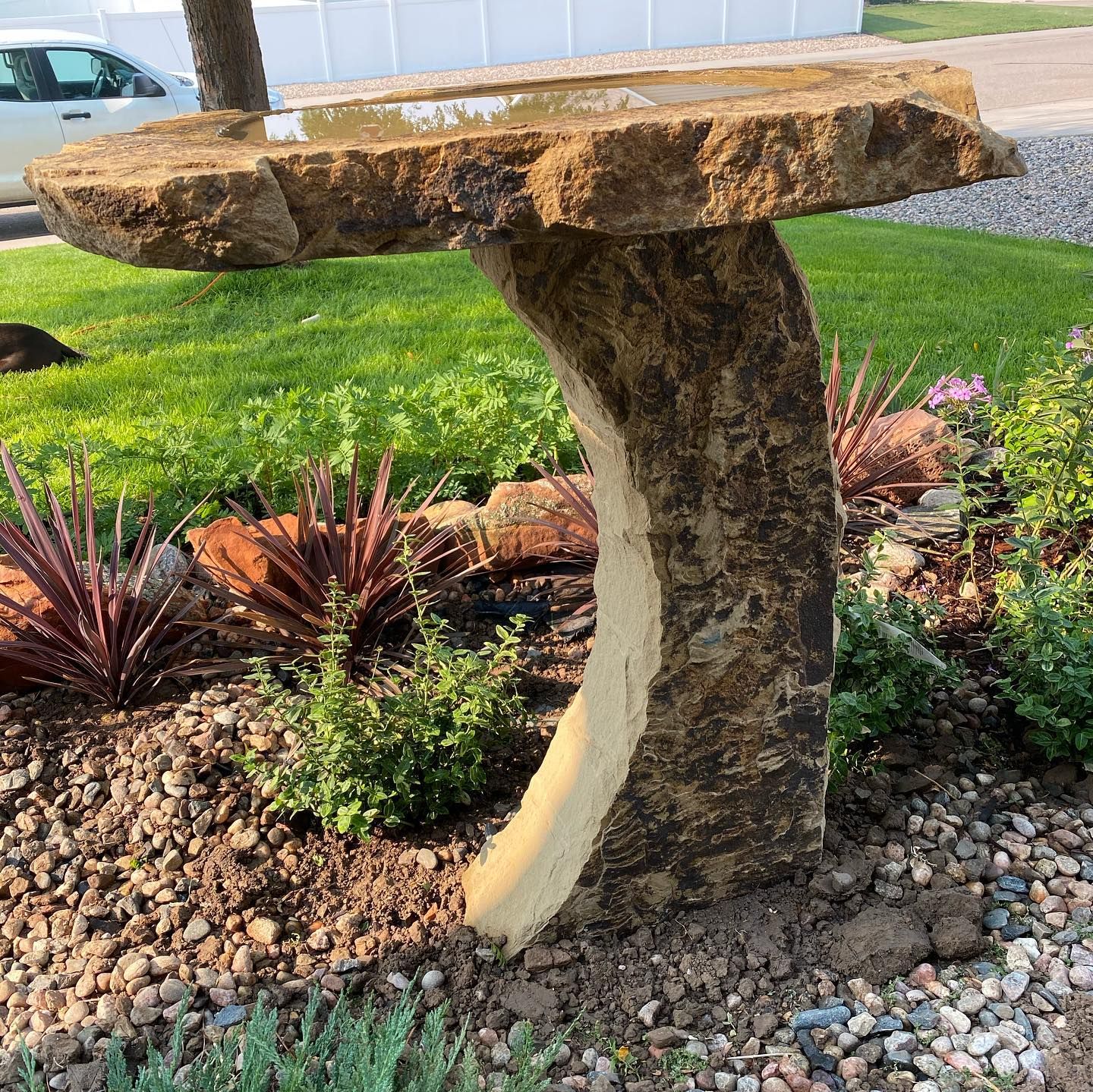 Rustic stone birdbath sculpture in a landscaped garden with rocks, grass, and plants.