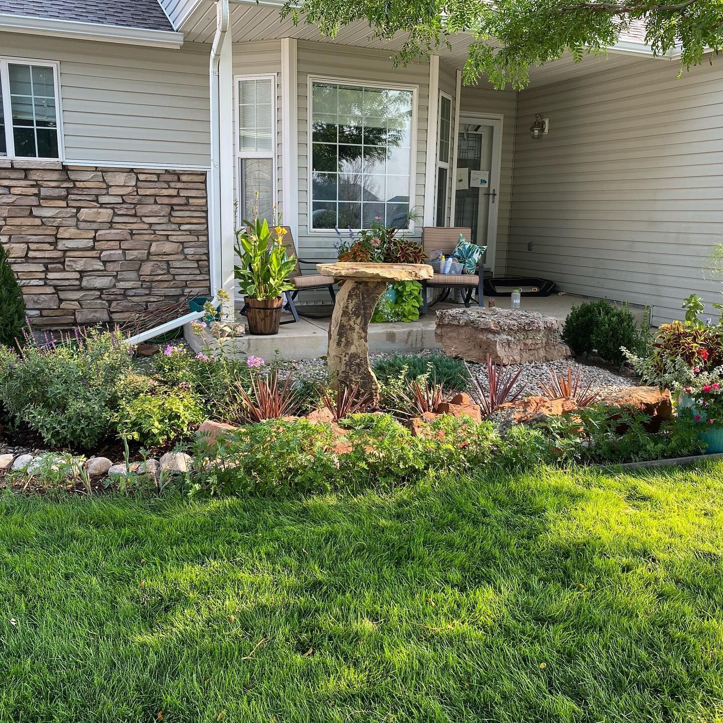 Front porch garden with flowers, stone edging, and a small birdbath beside a house.