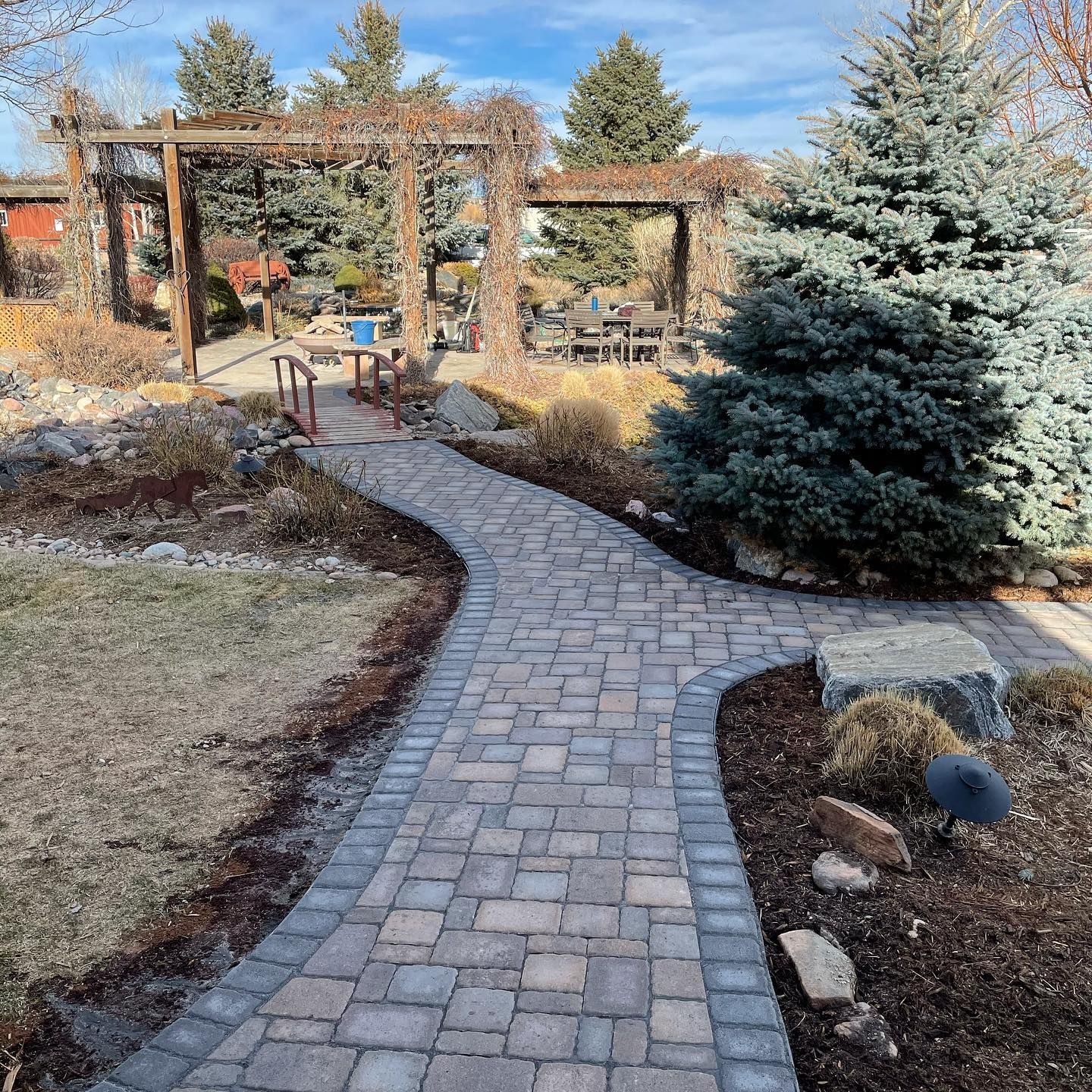 Curved brick garden path leading to a wooden pergola, with evergreen trees and landscaped shrubs.