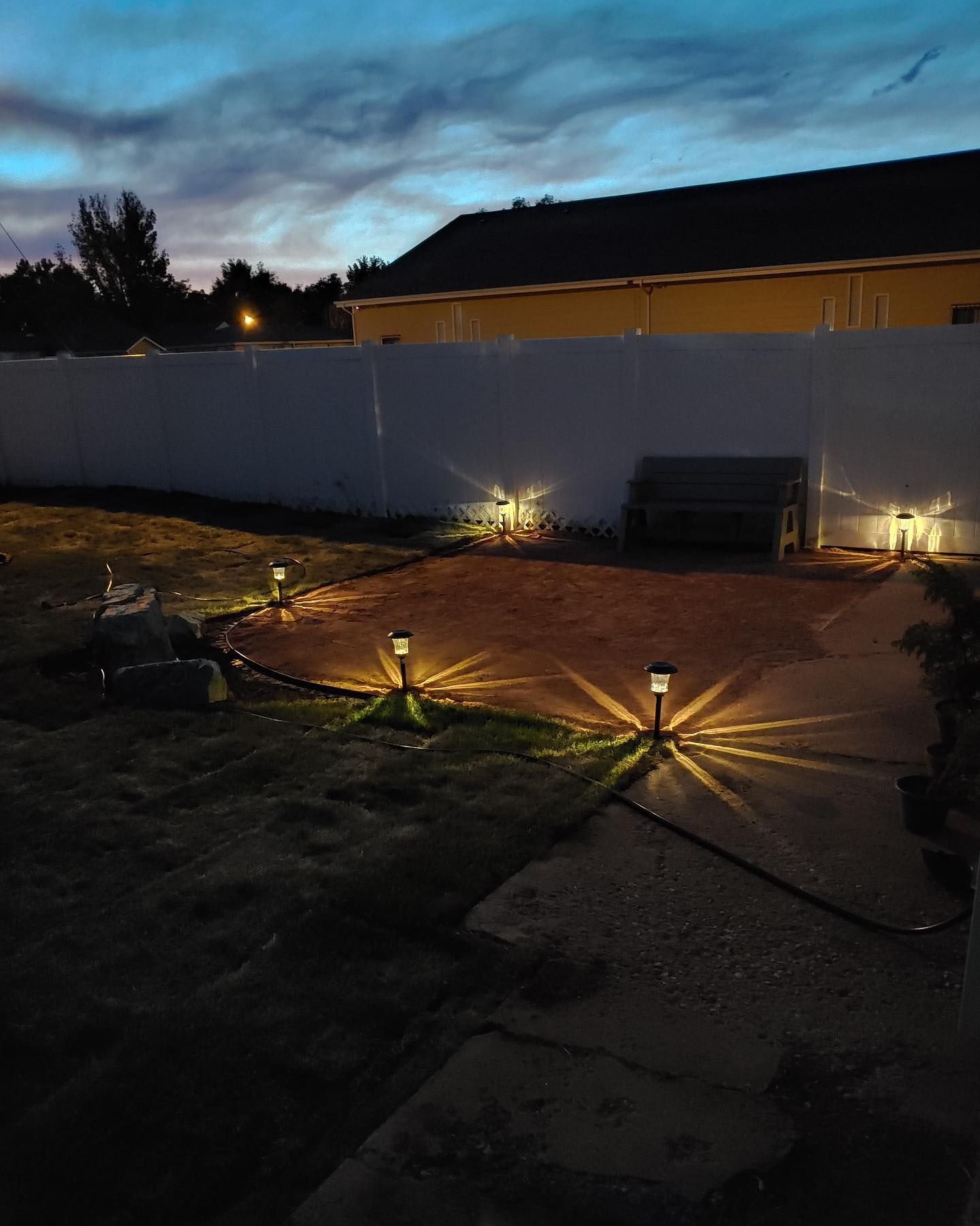 Backyard patio at dusk with path lights illuminating a paved area and white fence