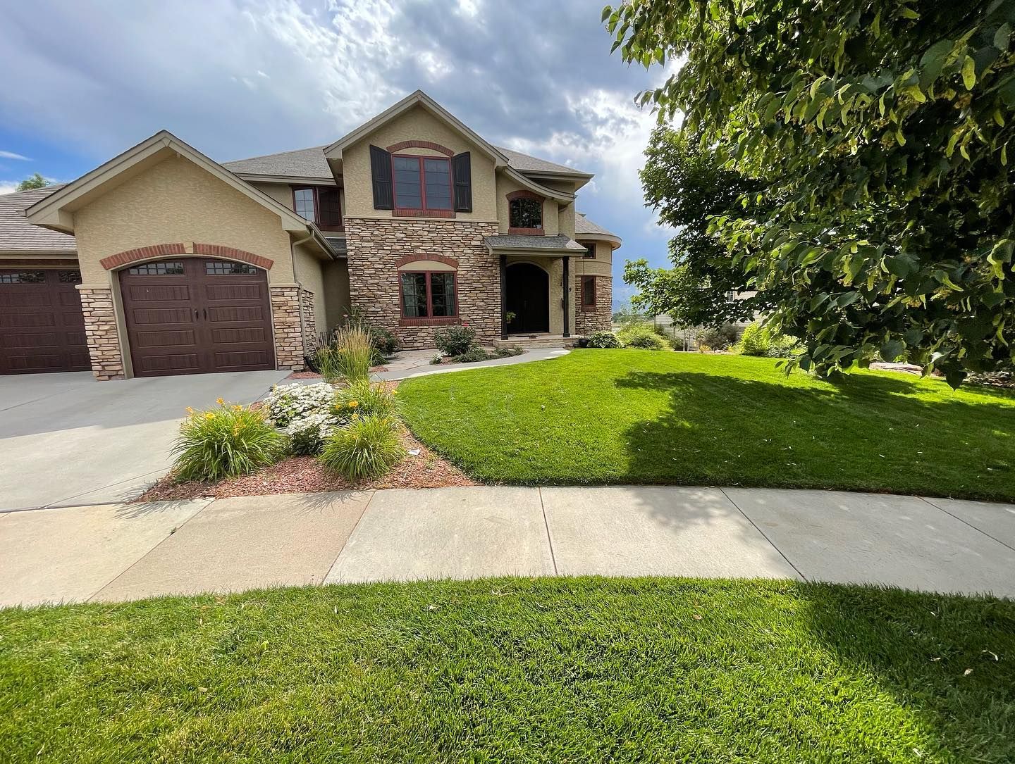 Suburban house with stone facade, two garages, and a green lawn under a cloudy sky