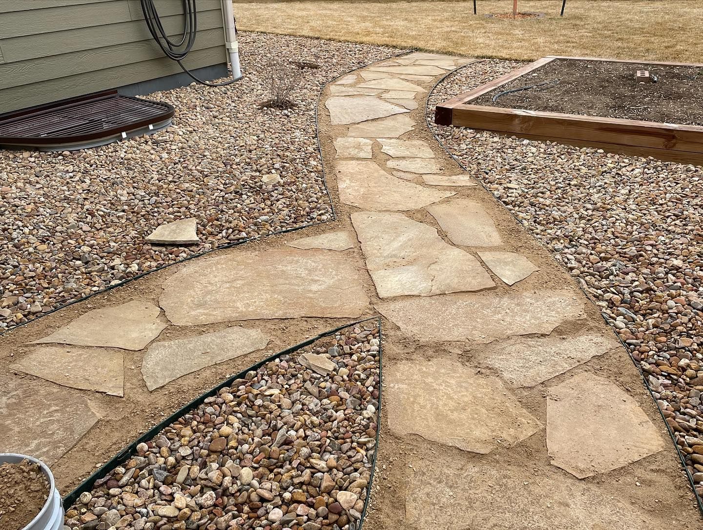 Stone garden walkway winding through tan gravel with a low raised bed on the right