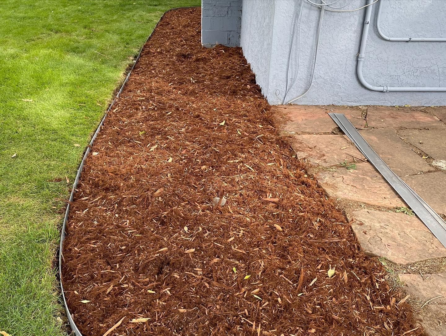 Fresh mulch bed edging a house beside a stone patio and green lawn.
