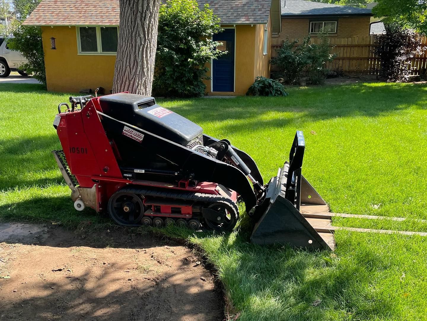 Red and black compact loader with bucket on grass in a yard near a house
