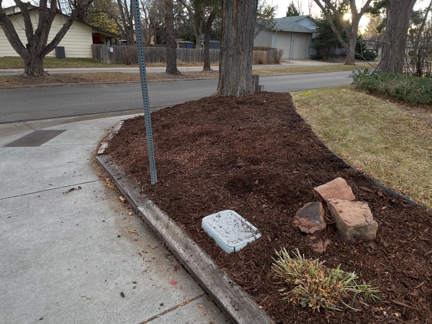 Mulched curbside planter beside a sidewalk, with a sign post and rocks near a residential street.