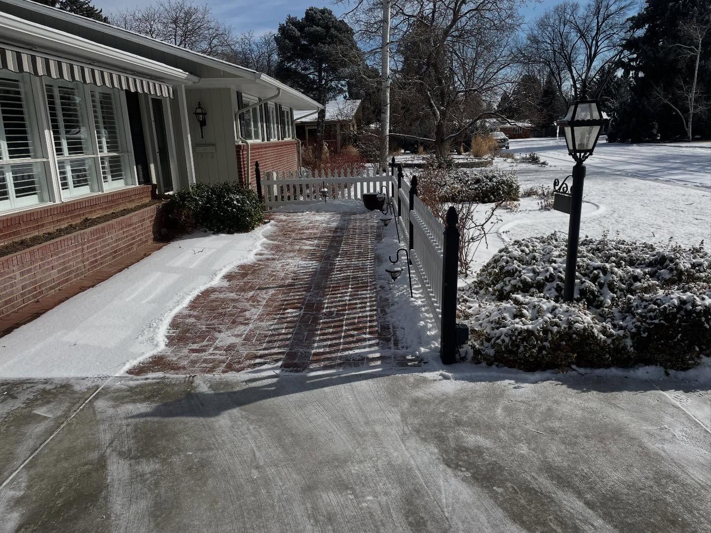 Snow-dusted brick walkway beside a house, leading to a front yard with trees and a lamp post.