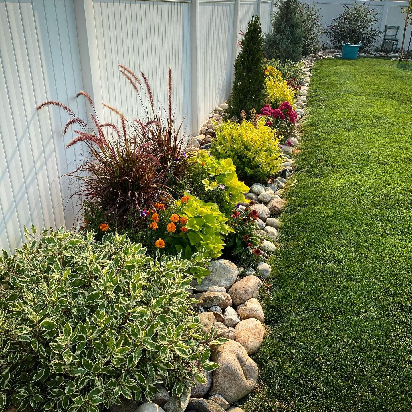 Colorful flower bed and ornamental grasses lining a white fence beside a green lawn