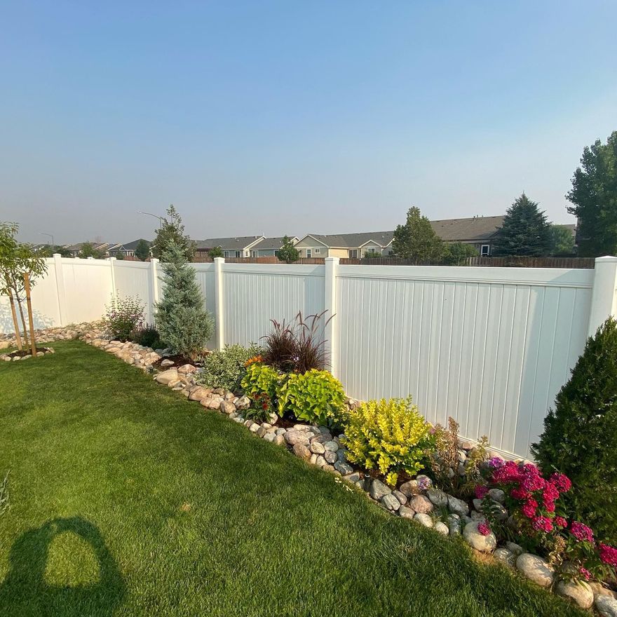 White vinyl fence borders a manicured lawn and colorful flower bed under a clear blue sky.