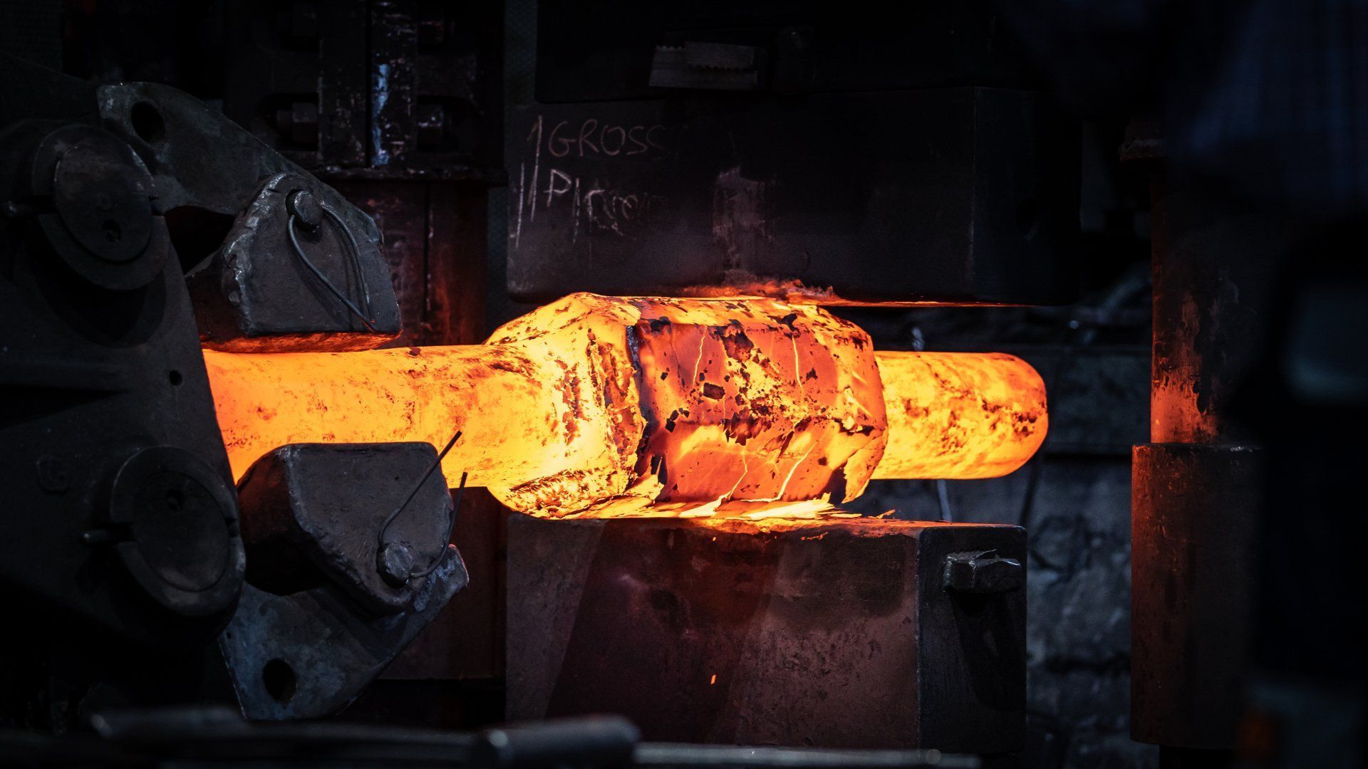 In un'officina buia, tra scintille e macchinari, viene forgiata una barra di metallo incandescente di colore arancione.