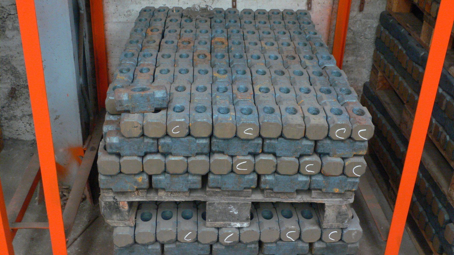 Pile di componenti metallici cilindrici grigi disposti in file su un pallet, incorniciati da travi arancioni.