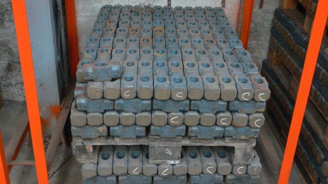 Parti o blocchi di metallo grigio impilati su un pallet all'interno di un'area di stoccaggio industriale, delimitati da binari arancioni.