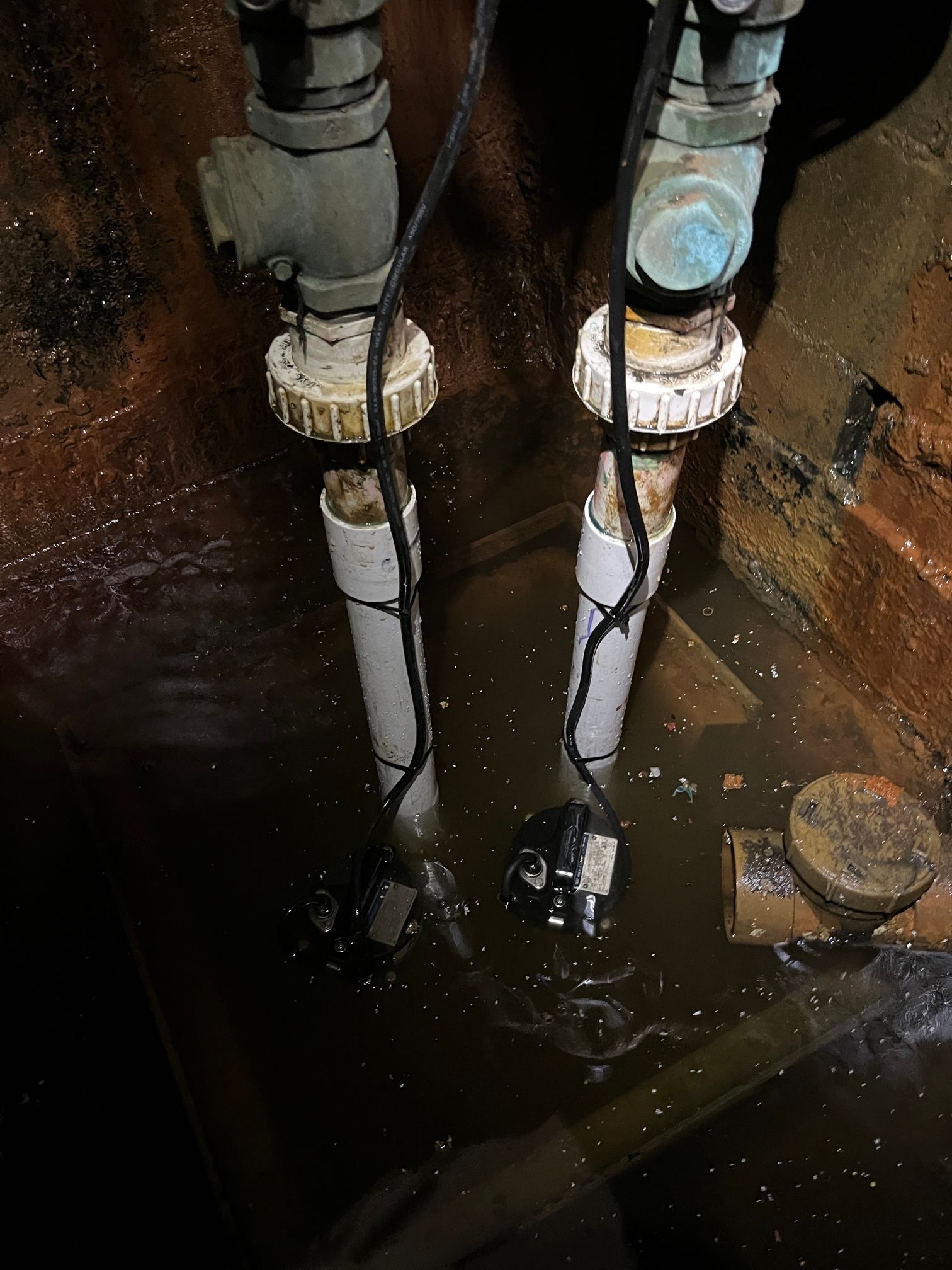 Two sump pumps submerged in water in a dark, brick-lined pit.