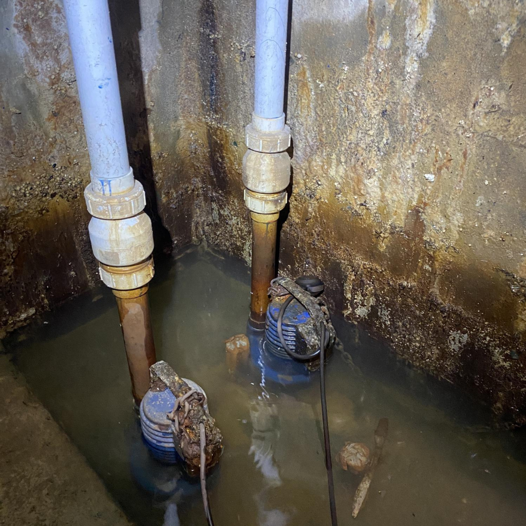 Sump pit with water, pipes, and submersible pumps in a concrete enclosure.