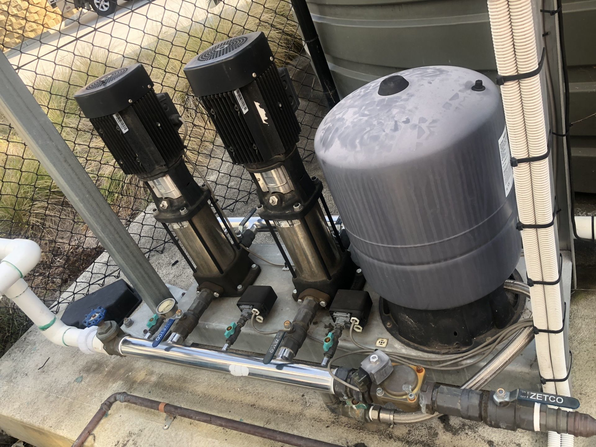 Three water pumps and a pressure tank, pipes, and wiring on a concrete base.