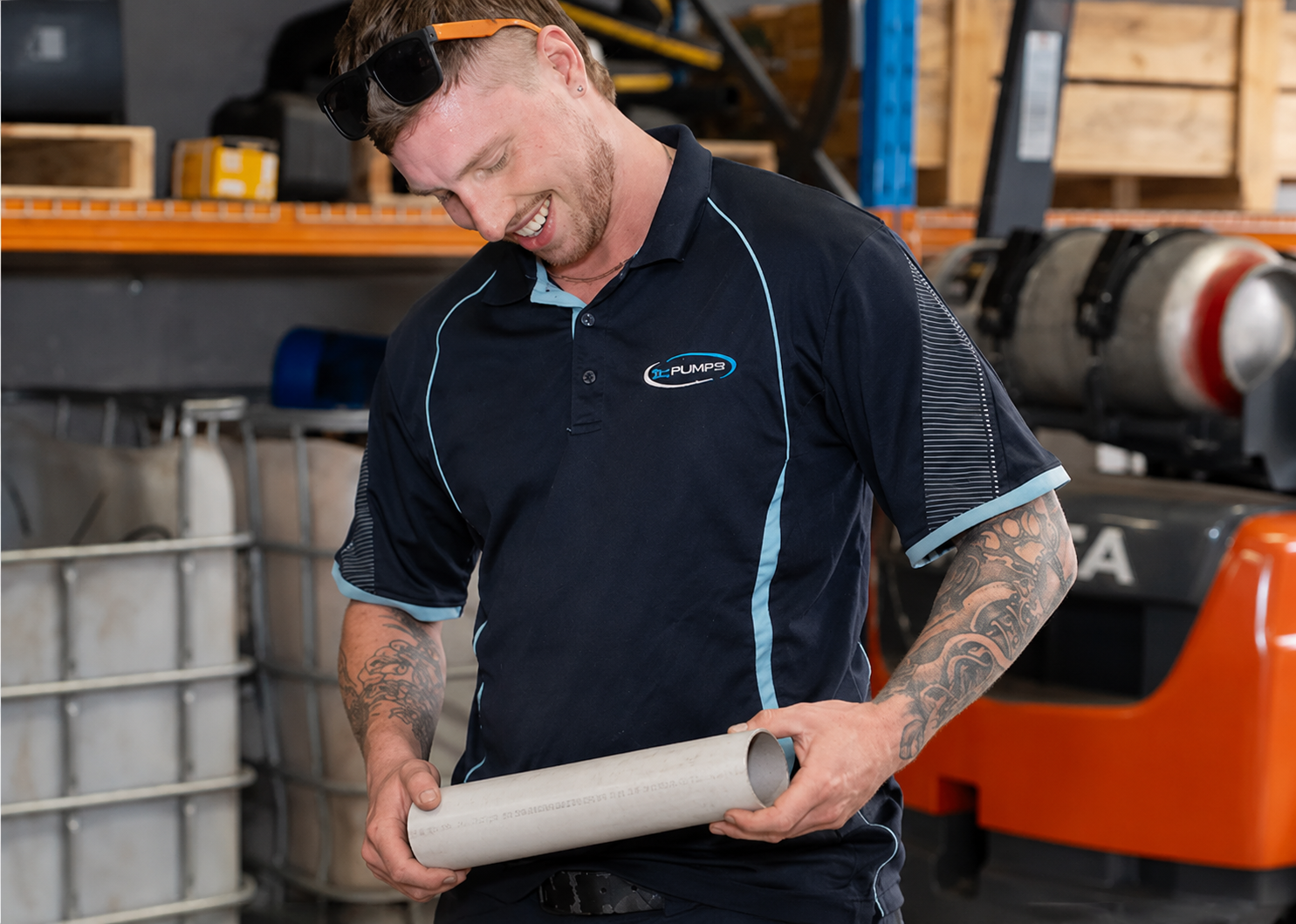 Worker in a workshop smiling while holding a white cylindrical plumbing part near machinery.