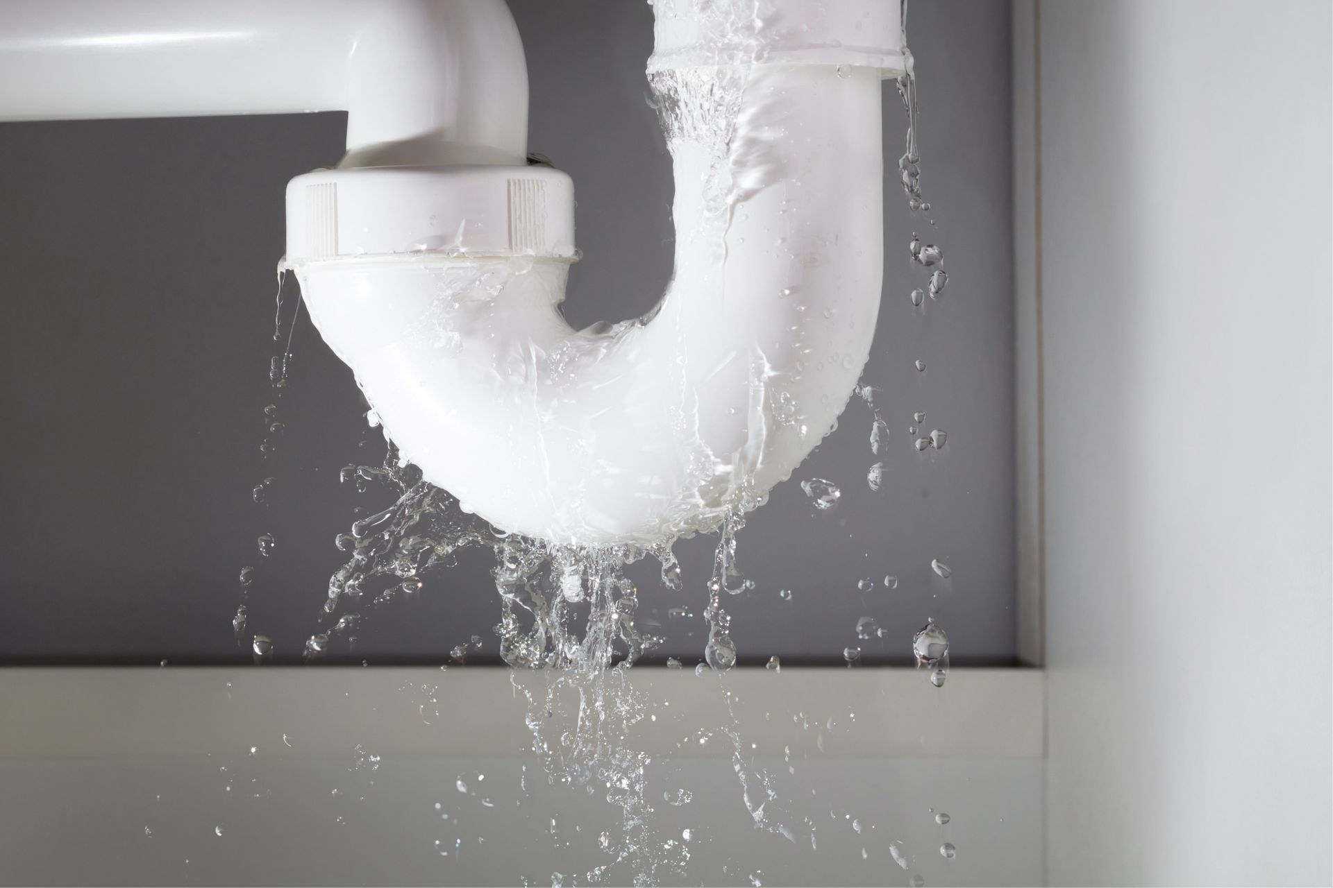 Leaking white plumbing pipe, water spraying downward, visible connection point.