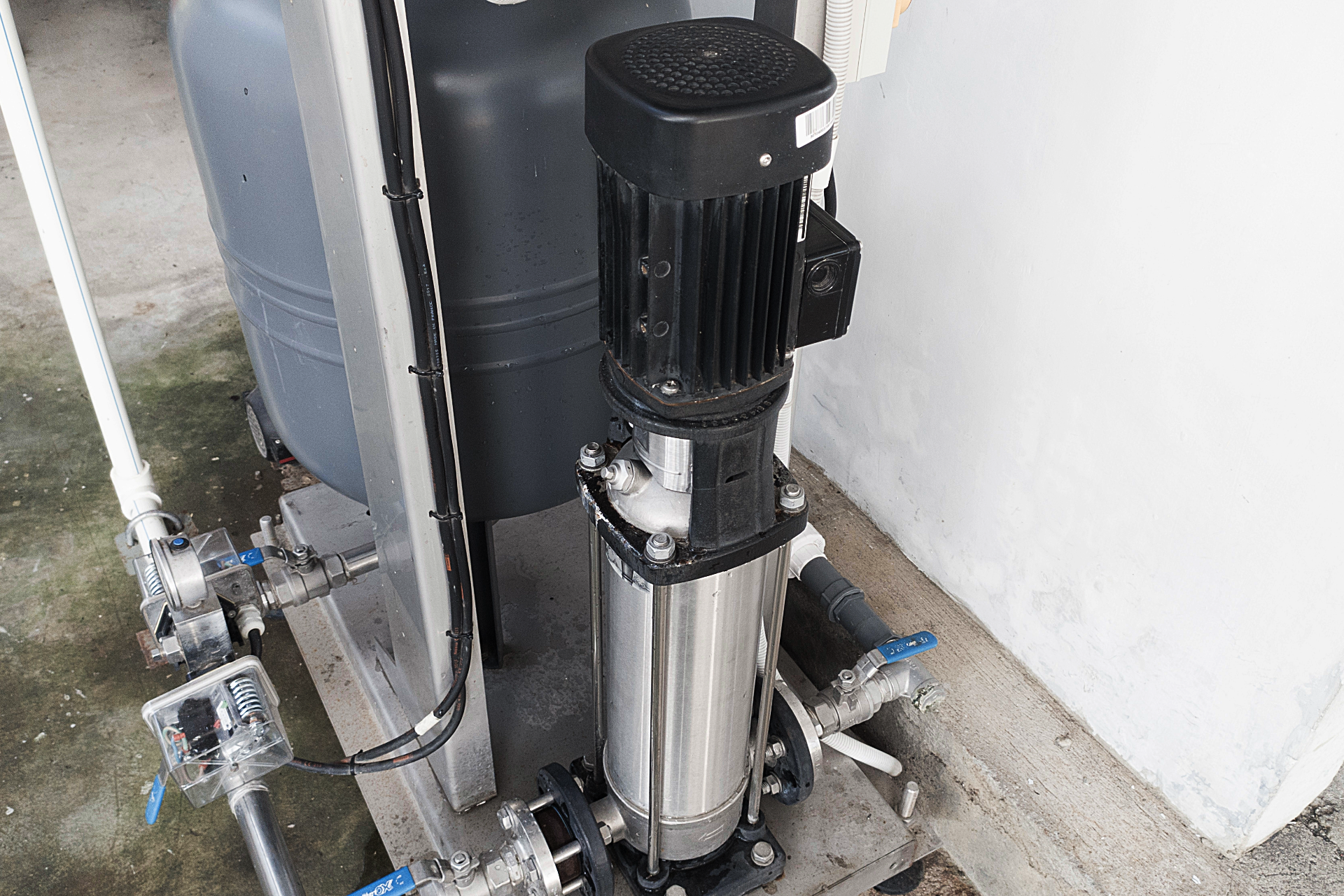 Vertical industrial water pump beside a large gray tank against a white wall outdoors