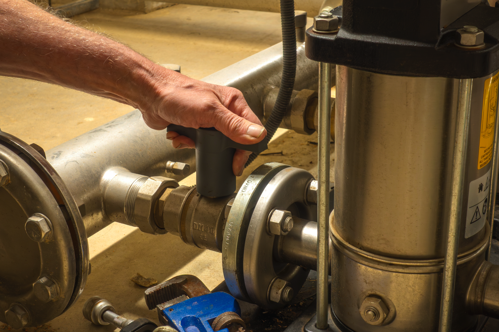 Hand holding a black tool on a metal pipe valve in an industrial setting