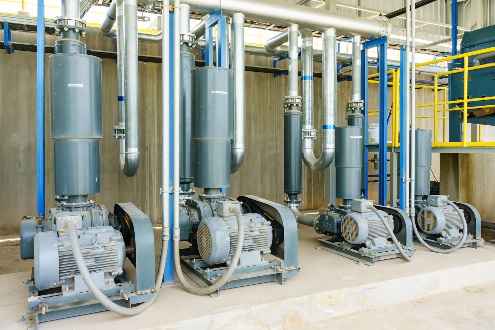 Gray industrial blowers with silver piping in a factory setting.