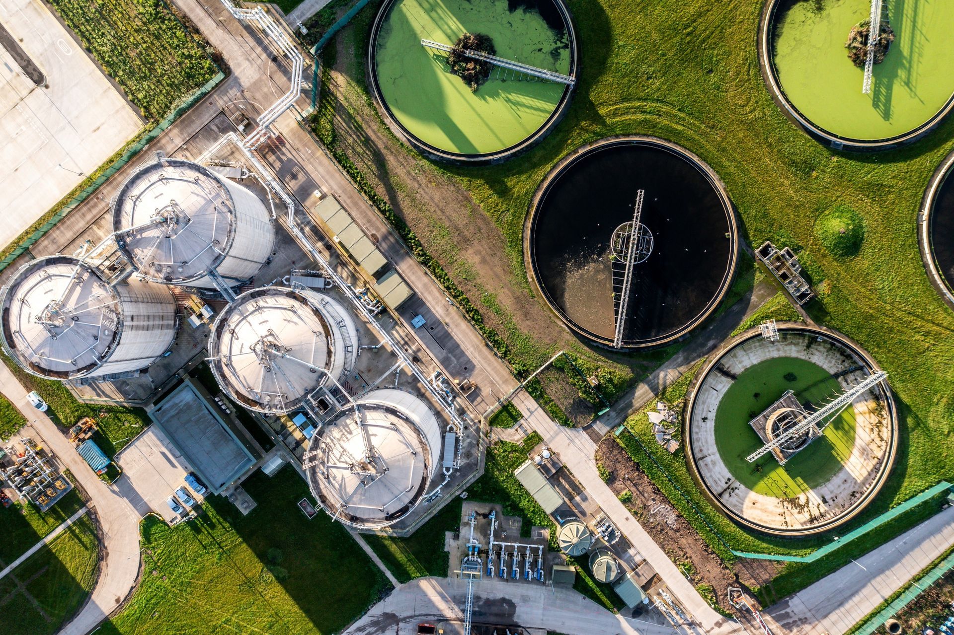 Aerial view of a wastewater treatment plant with several tanks and circular basins, various colors, and green surroundings.