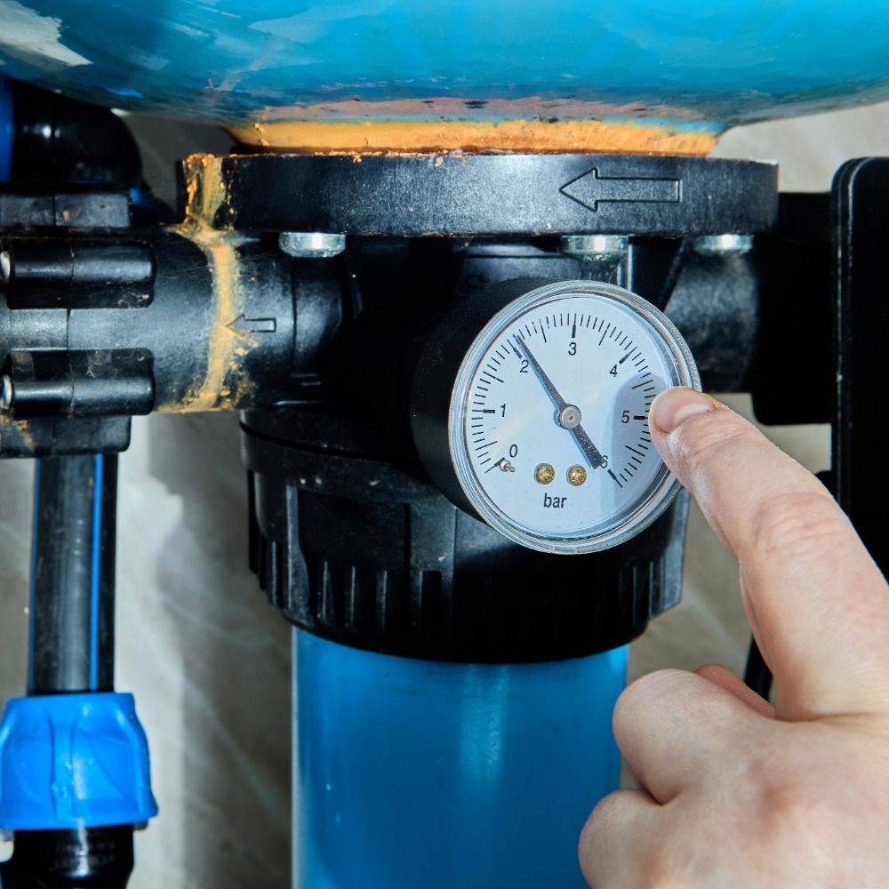 Hand pointing at pressure gauge on a water filtration system. The gauge reads near 2 bar.