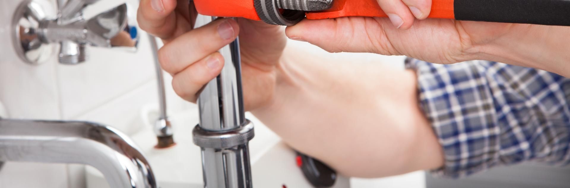 A pair of hands using a pipe wrench to tighten a metal plumbing pipe beneath a sink.