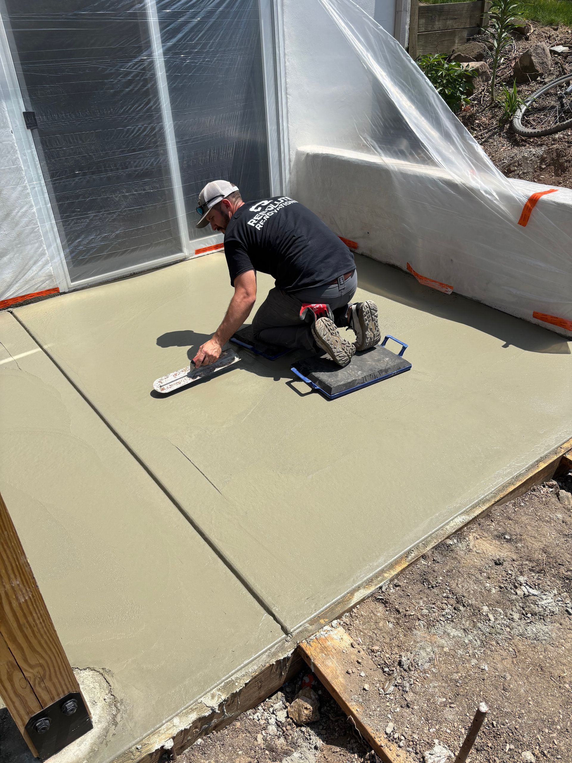 A construction worker smoothing fresh concrete on a patio. He kneels, using a trowel.