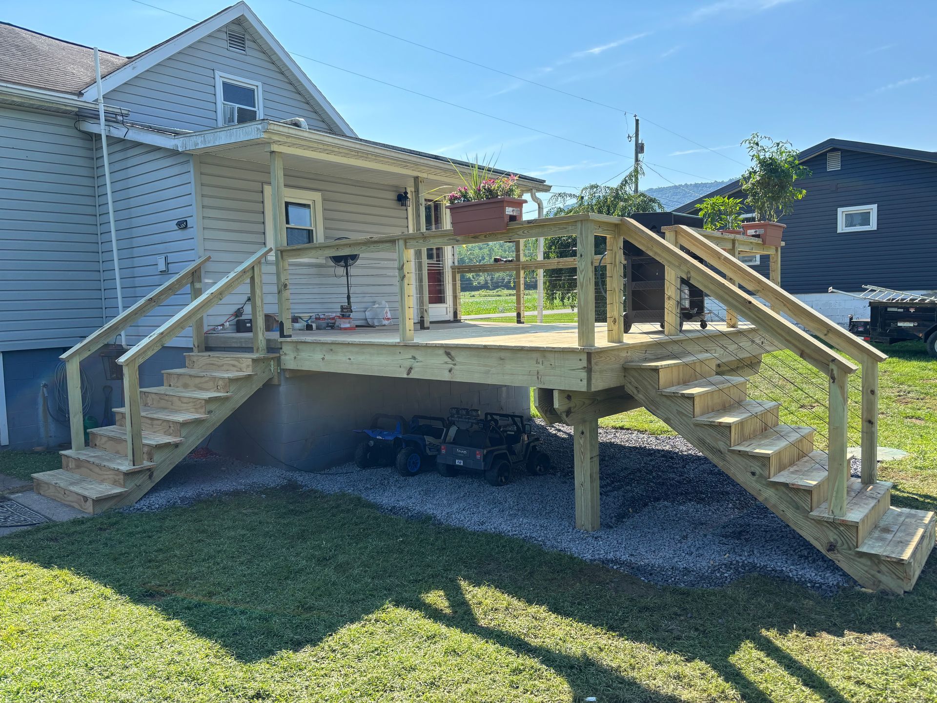 A new wooden deck attached to the side of a light gray house, featuring two sets of stairs leading up from a gravel area.