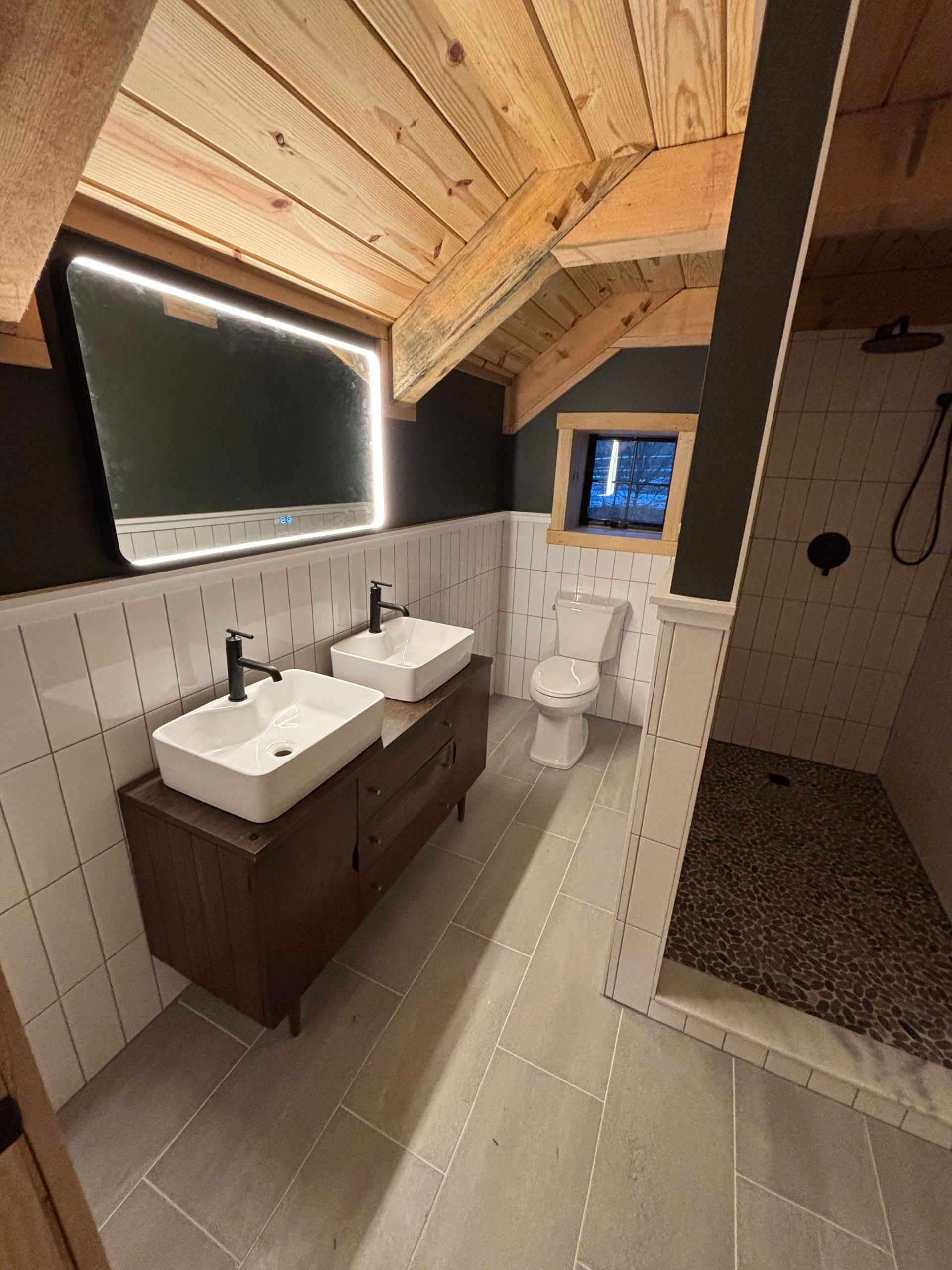 Bathroom with white tile, dark vanity, and a pebble-floored shower. Wood ceiling and black accent wall.