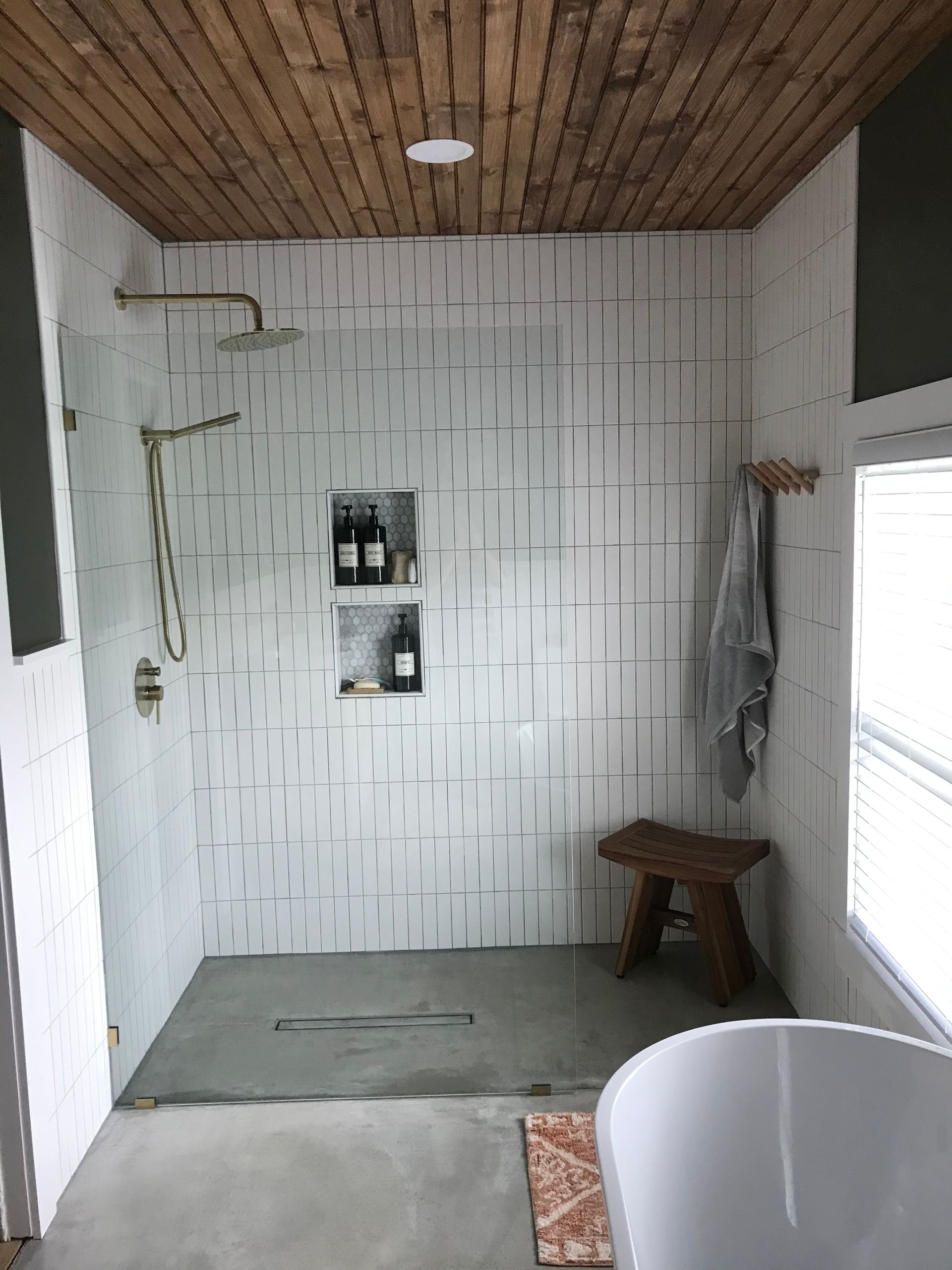 Bathroom with wood ceiling, white tile shower, two built-in shelves, and a wooden stool.