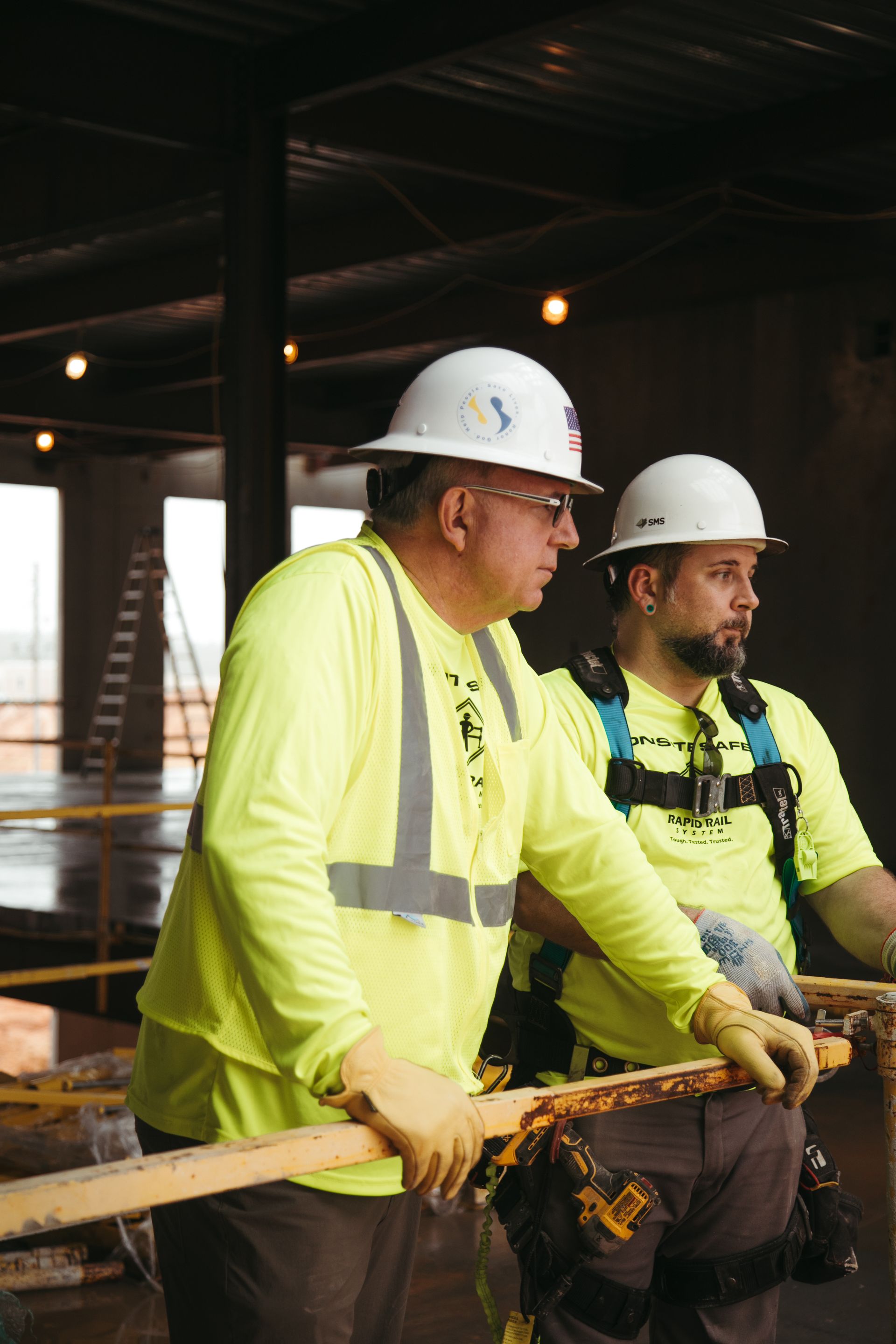 Two construction workers in hard hats and neon safety shirts at a worksite, leaning on a railing.