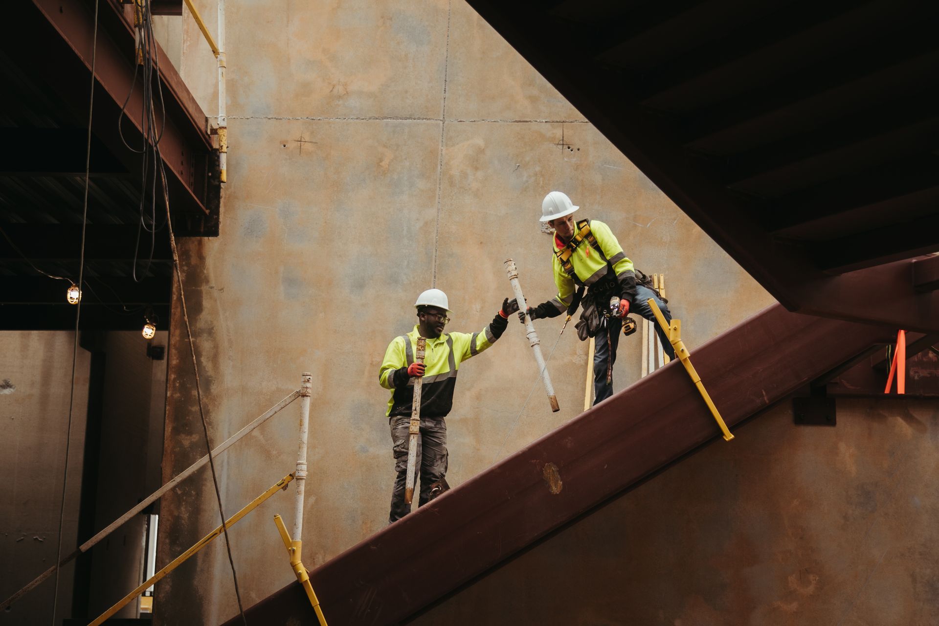 Two construction workers in safety gear stand on a sloped steel beam at a building site.