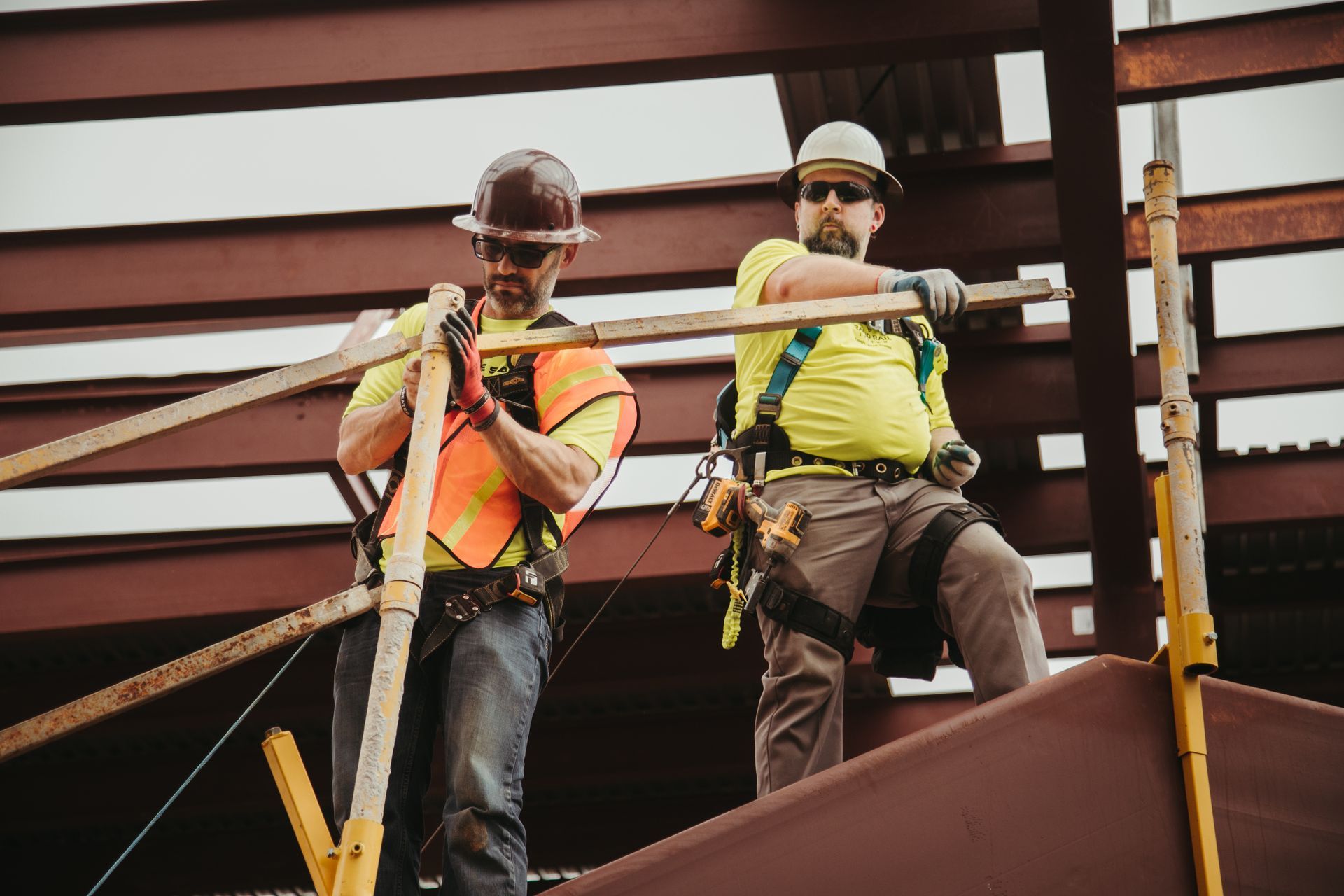 Two construction workers in hard hats and safety vests working on a steel beam.