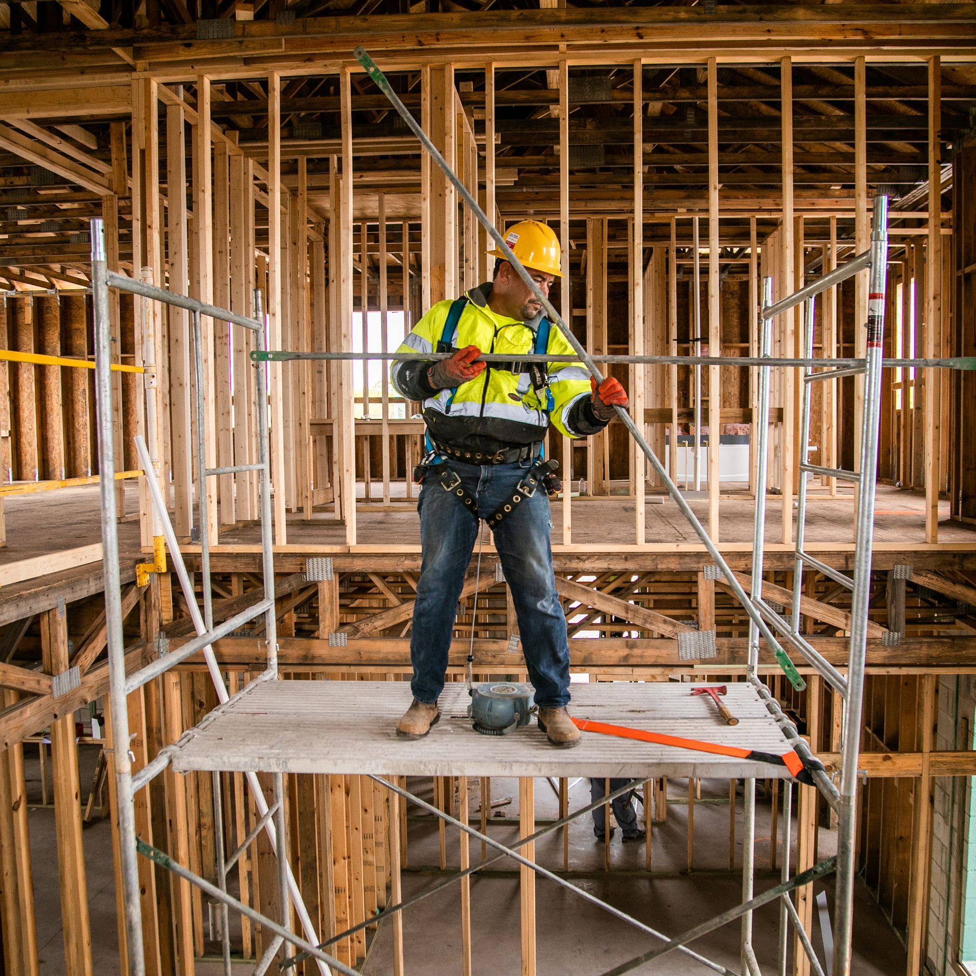 Construction worker in a hard hat and reflective vest on scaffolding inside a wooden house frame.