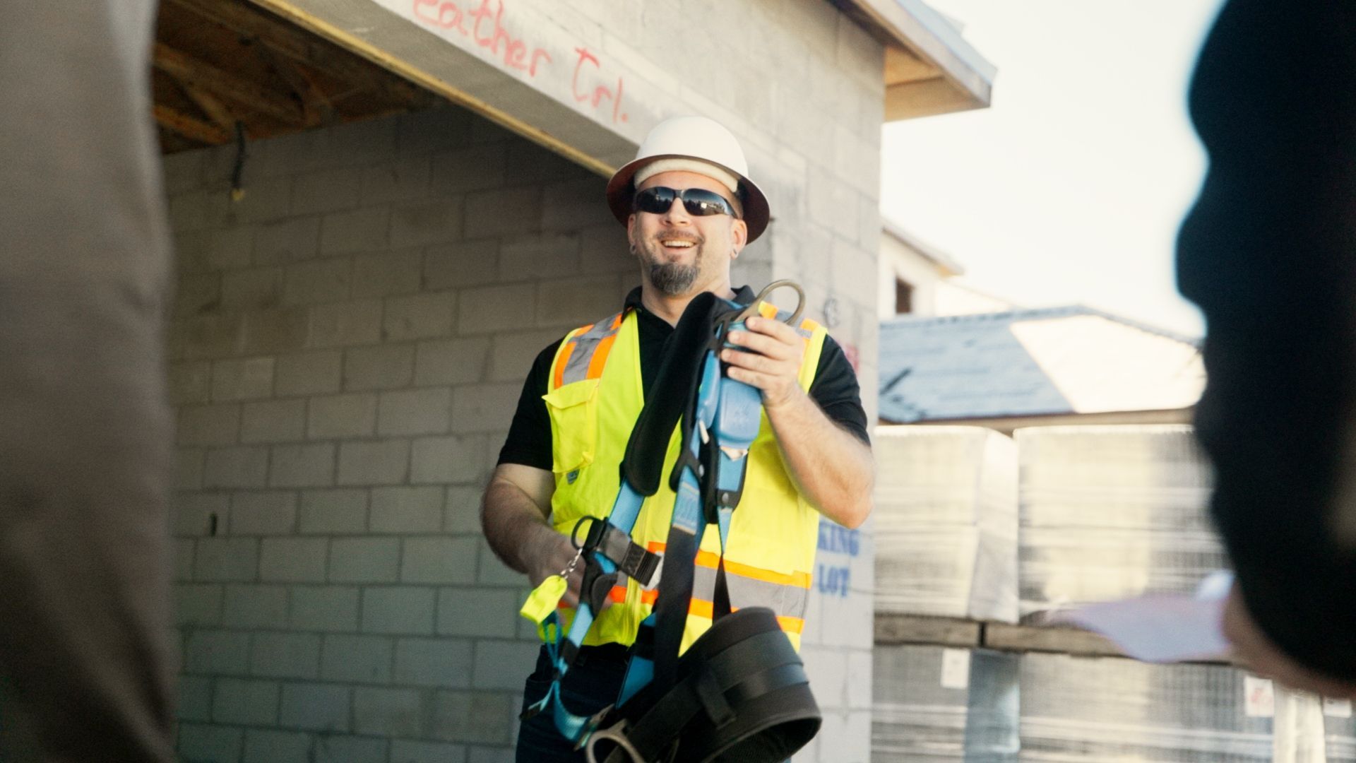 Worker in a neon safety vest and hard hat smiles while holding tools at a construction site.