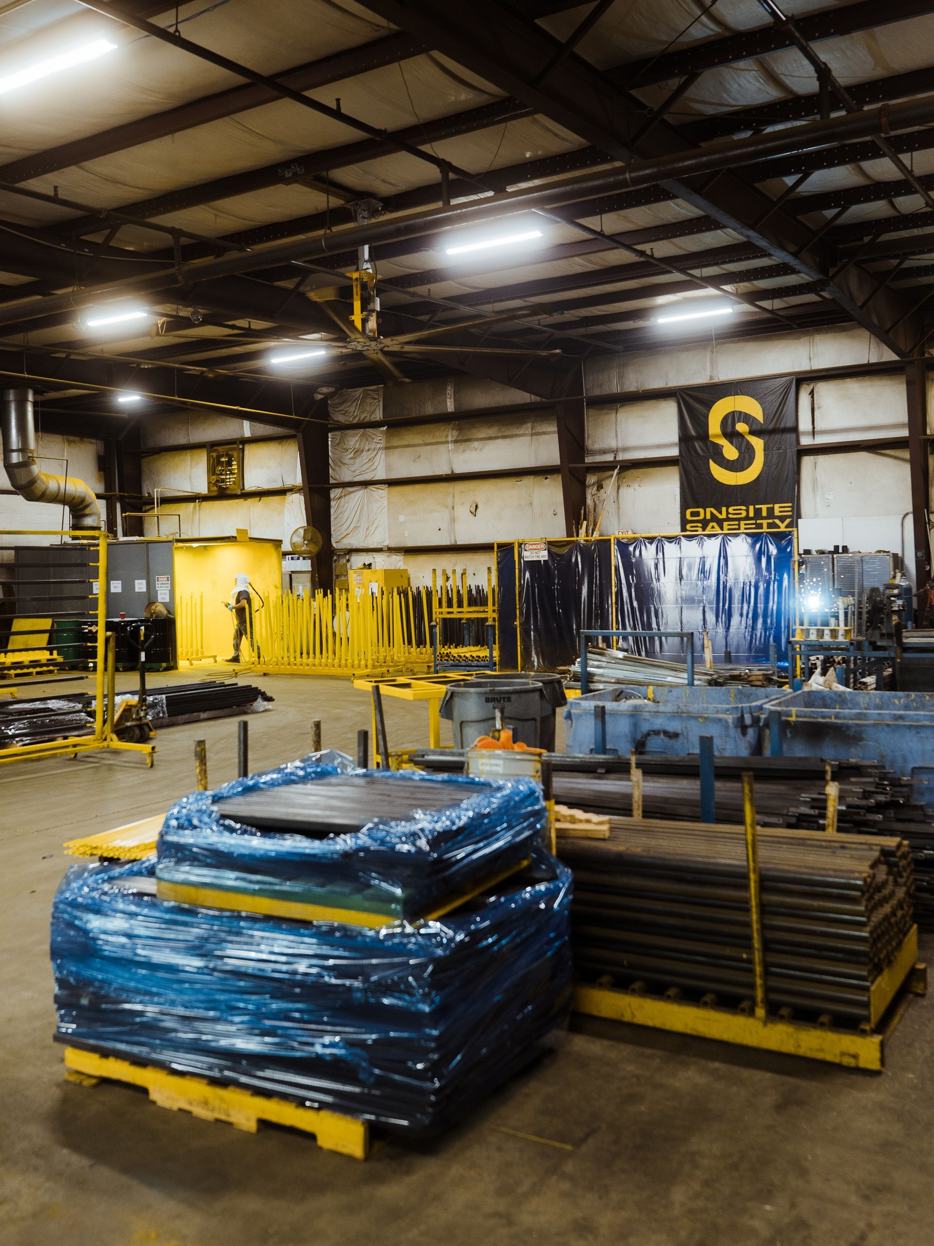 Industrial warehouse with stacked pallets, blue-wrapped materials, and yellow equipment under bright overhead lights