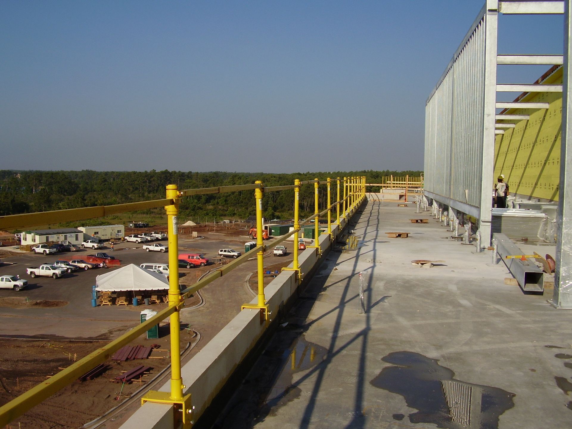 Rooftop construction walkway with yellow railings beside a tall wall, overlooking a parking lot and trees.