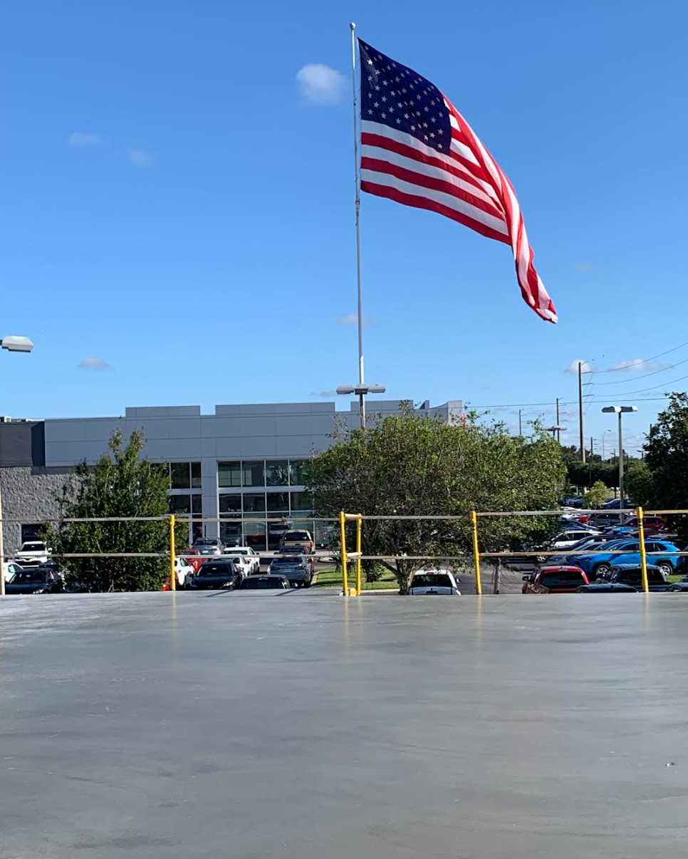 Large American flag waving over a parking lot beside a low commercial building on a sunny day