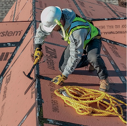 Worker in hard hat and safety vest securing roofing materials on a sloped roof with tools and coiled rope.