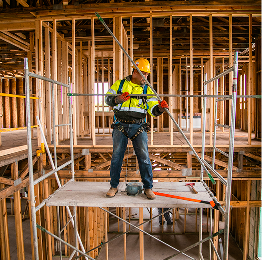 Construction worker in yellow vest and hard hat on scaffolding inside a wooden house frame.