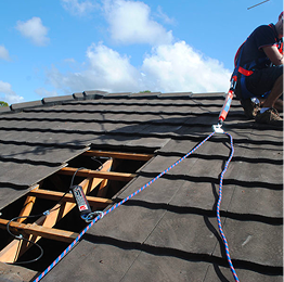 Roofer on a shingled roof repairing a damaged section with exposed wooden framing and safety ropes