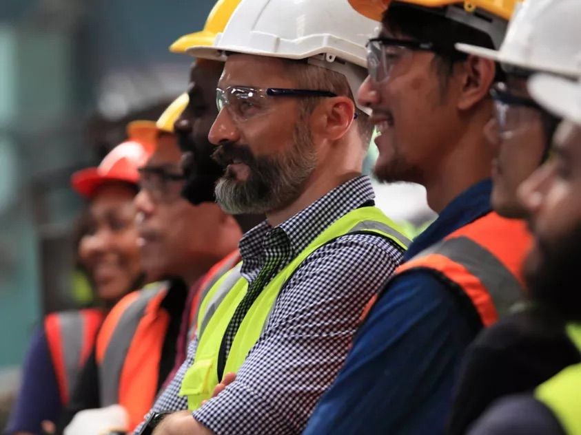 Workers in hard hats and safety vests standing in a workshop, listening attentively.