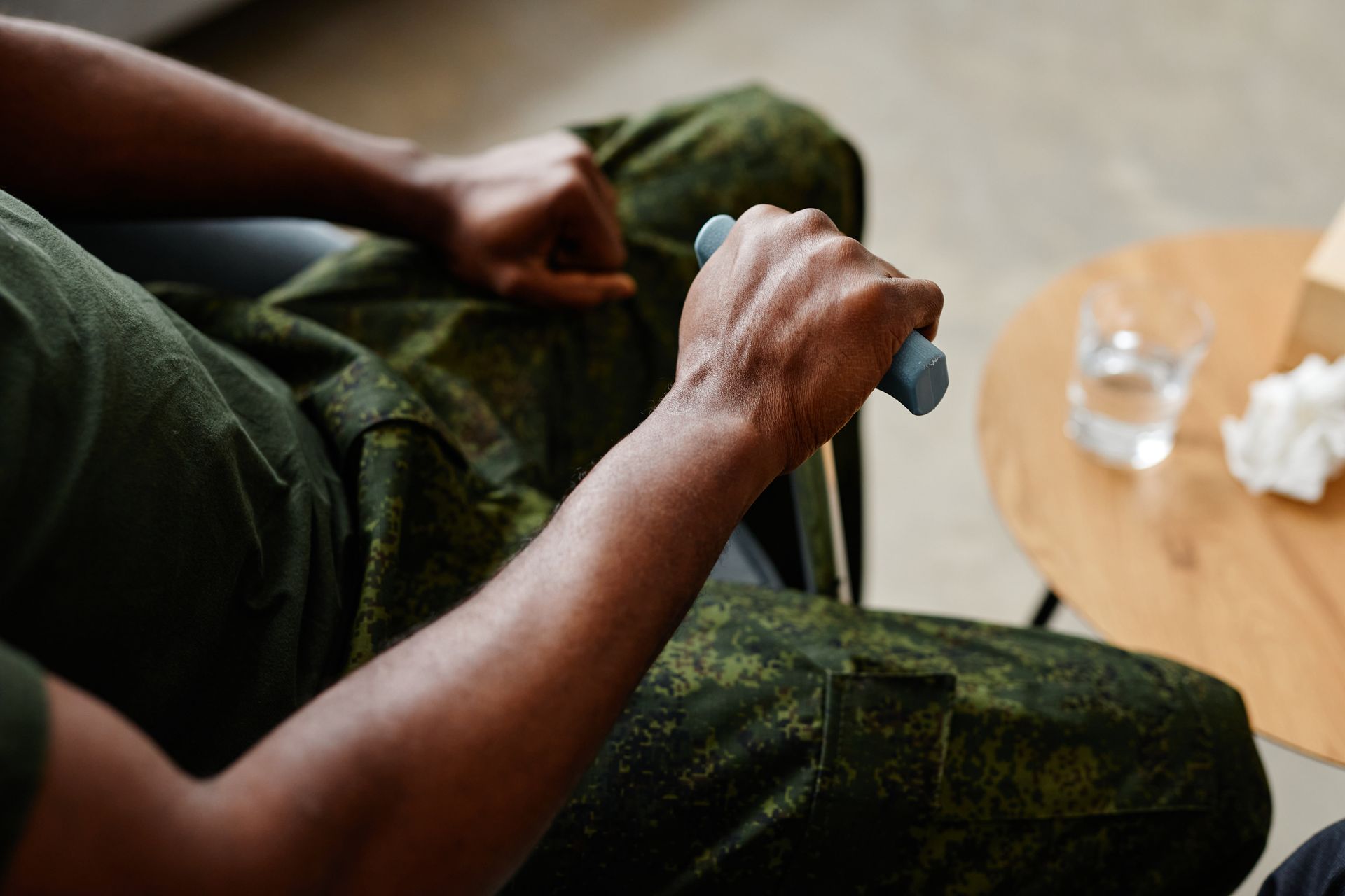 Person in military fatigues, gripping a blue walking cane, seated in a chair. Beside him are a glass of water and tissues.