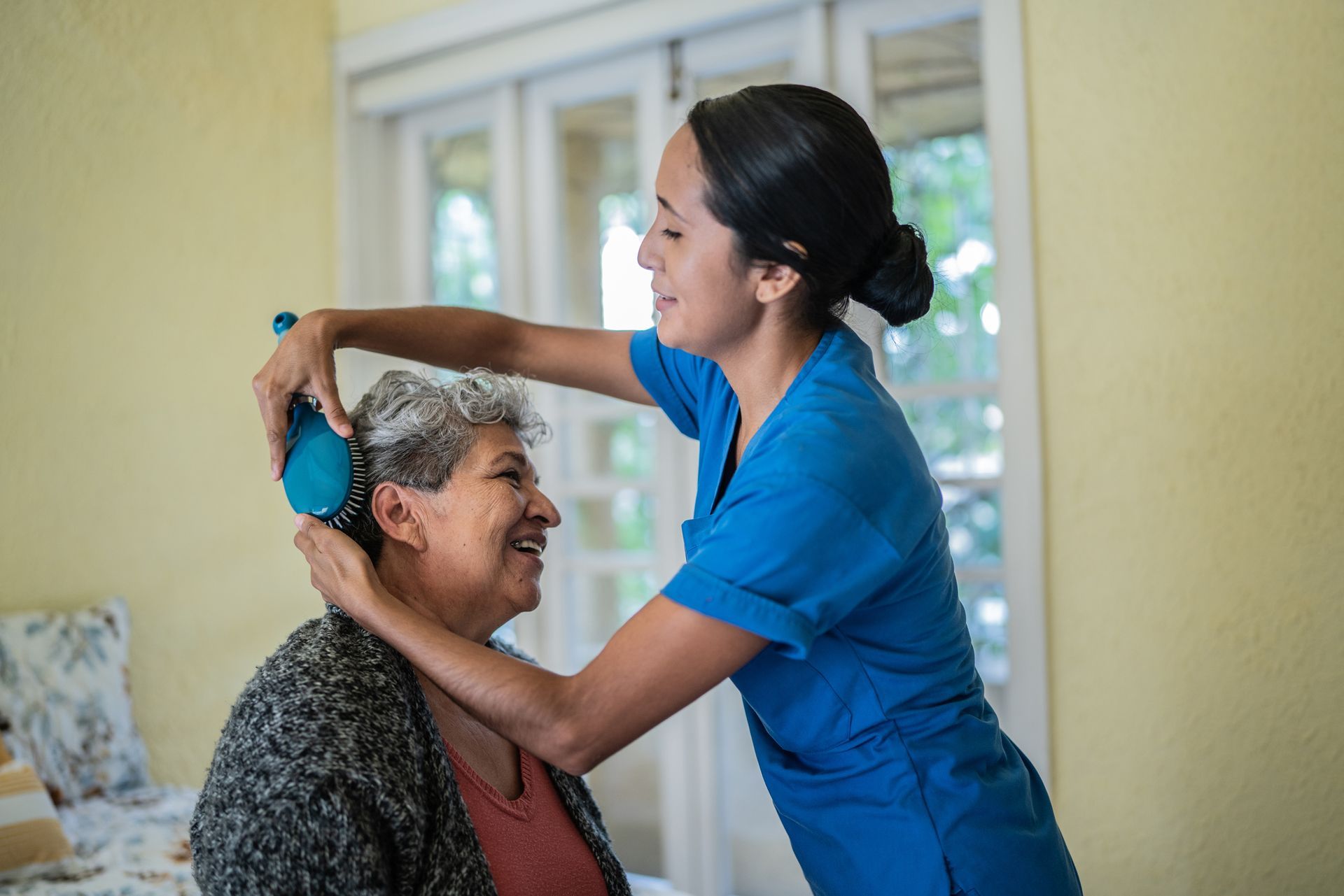 A young woman brushes an elderly woman's hair. Both smile in a yellow-walled room.