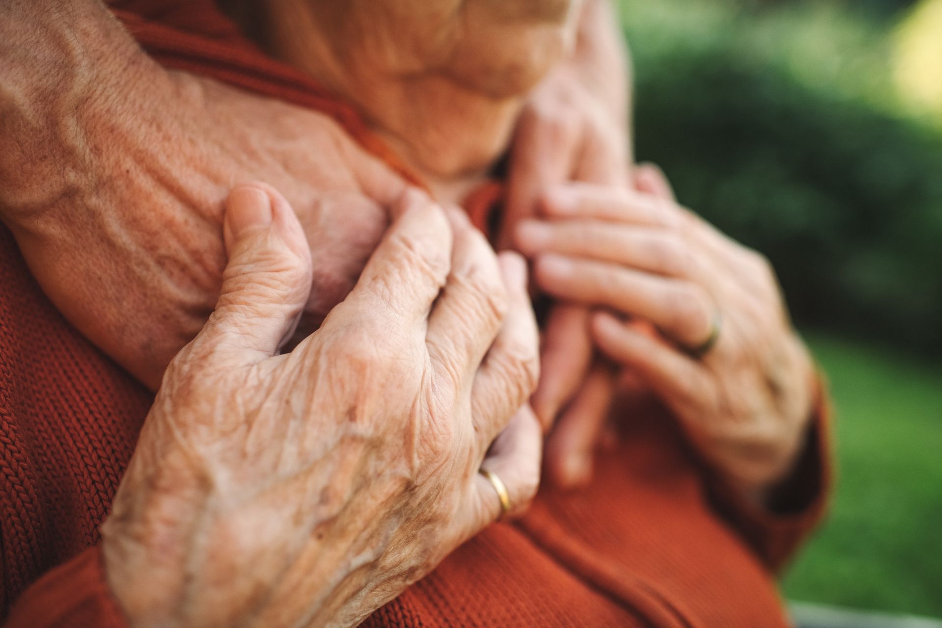 Two elderly people's hands clasped in a comforting embrace. They are wearing orange sweaters and gold rings.